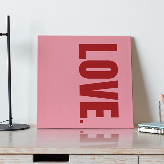 Pink 'LOVE' sign on a wooden desk with a black lamp and coffee cup.